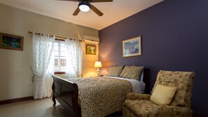 Un dormitorio con cama, una silla, ventana con cortinas, lámpara y dos cuadros en la pared.