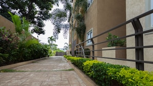 A paved pathway flanked by lush greenery and a building with a beige facade and metal railings.