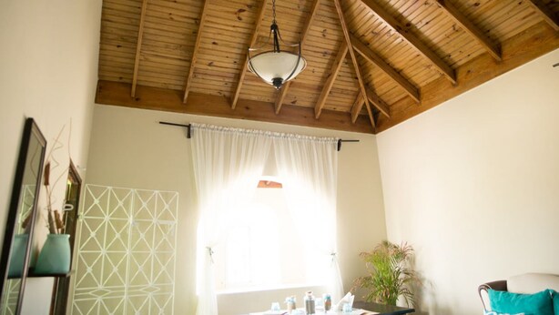 A room with a wooden ceiling, a hanging light fixture, a window with white curtains, a mirror, a geometric wall decoration, and a potted plant. - Apartment in Hellshire near Beach (Hellshire)