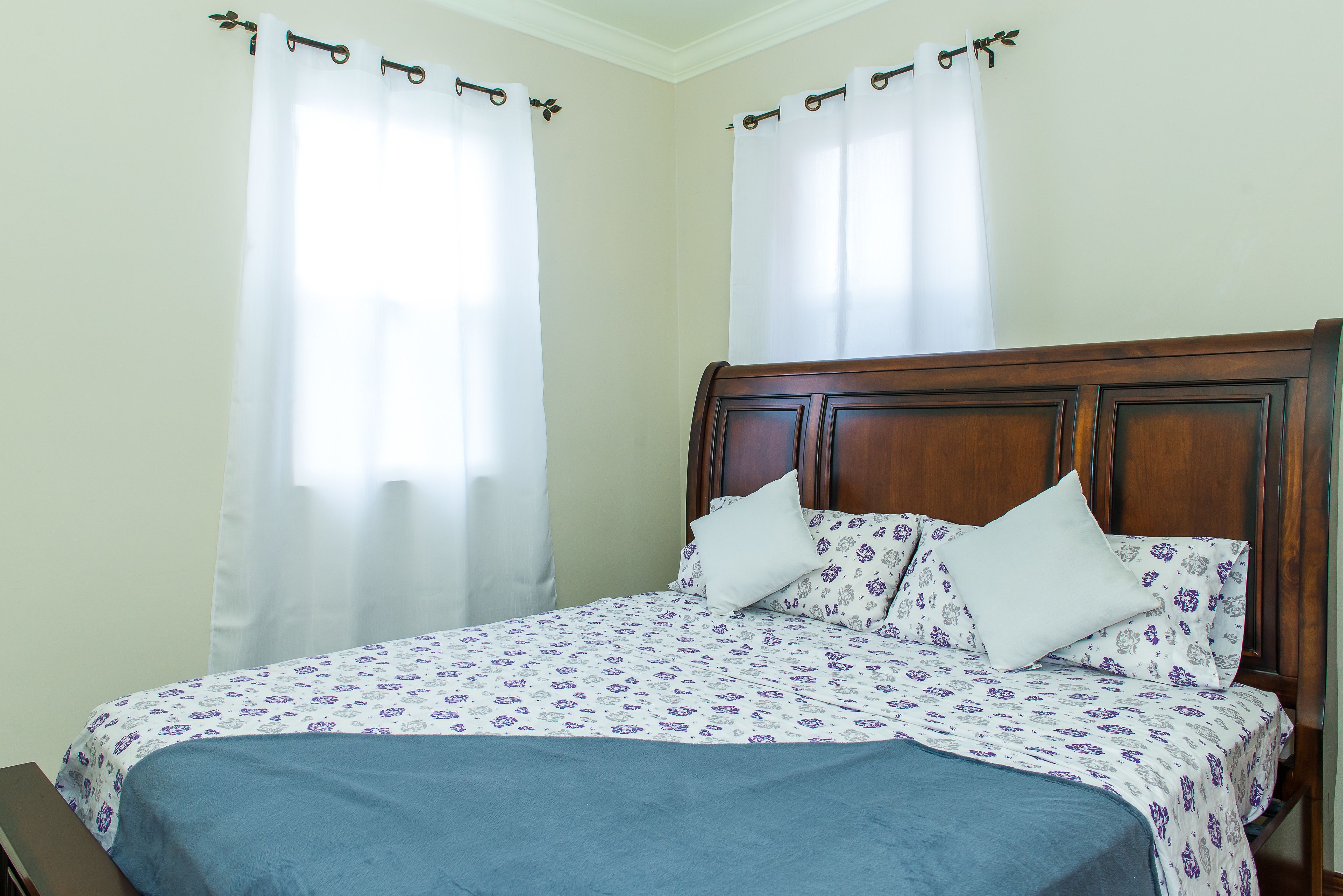 A neatly made bed with a floral bedspread, a wooden headboard, and white curtains.