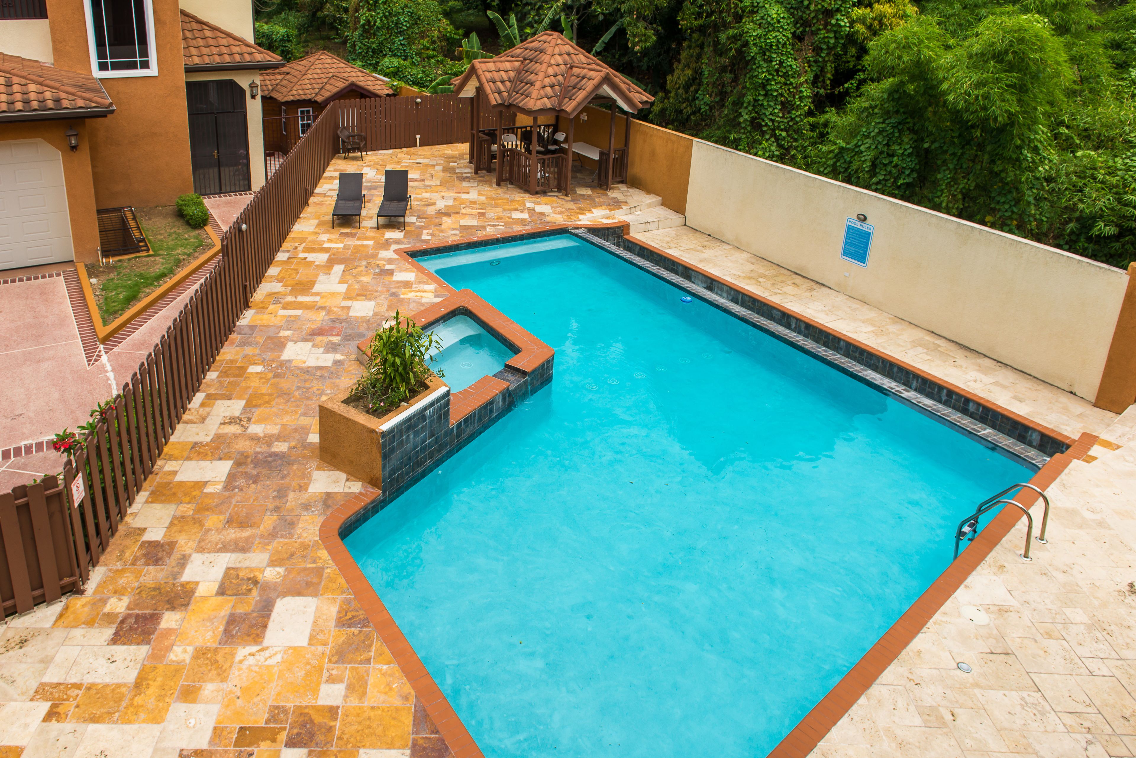 A rectangular swimming pool with a tiled deck, surrounded by a wooden fence and a small garden area.