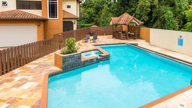 Een zwembad met een betegeld zonnedek, een kleine jacuzzi en een houten terras met stoelen.