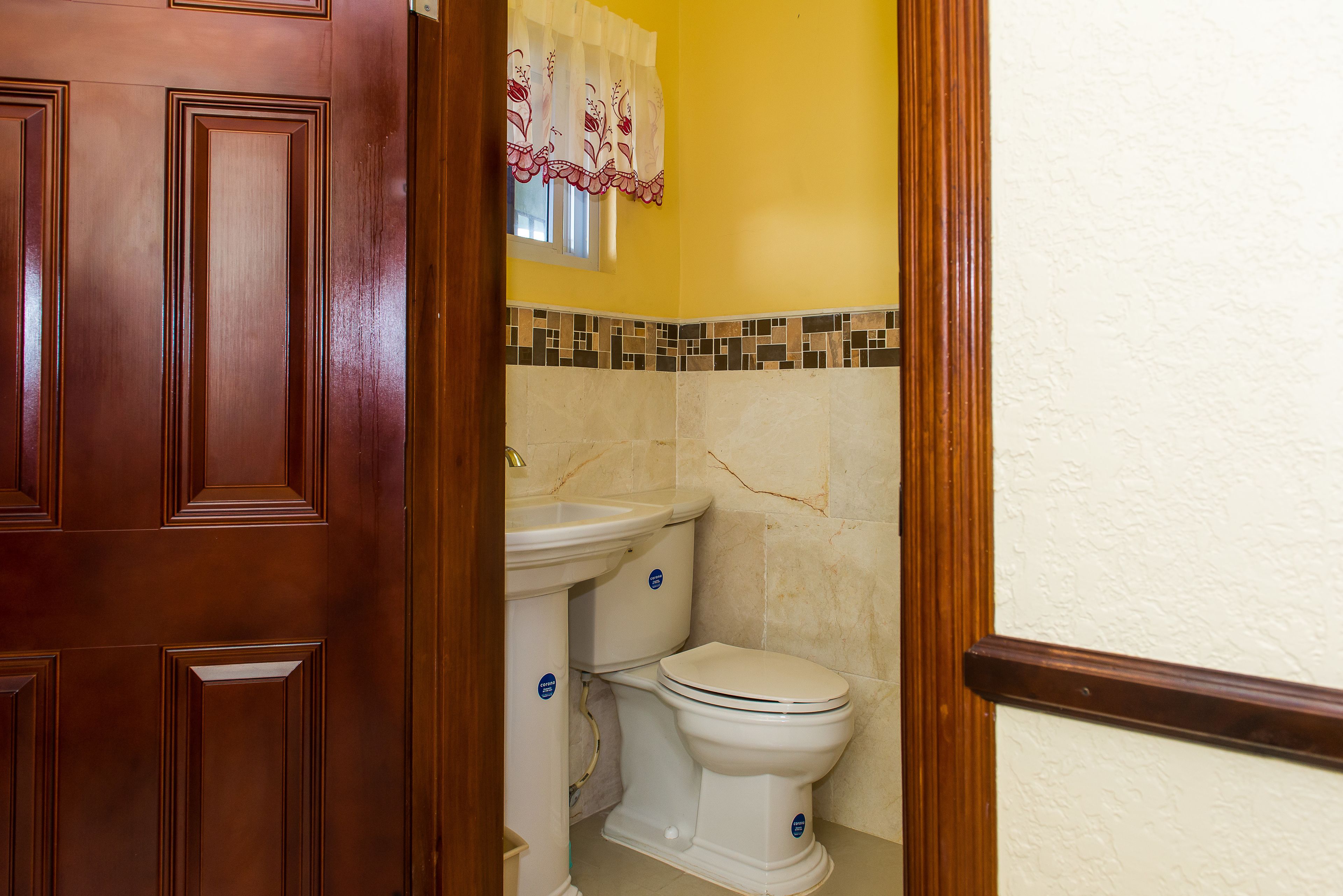 Un baño con puerta de madera, un inodoro, un lavamanos y una ventana con cortina floral.