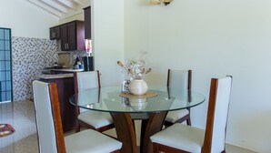 A dining area with a glass table surrounded by four chairs, a vase with flowers, and a kitchen in the background. - Ocho Rios Beach House at Dax Hall (Ocho Rios)