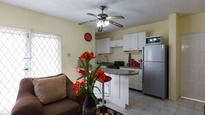 A living room with a brown sofa, a coffee table with a vase of flowers, a ceiling fan, and a kitchen area with white cabinets, a refrigerator, and a microwave. - Kgn Most Centrally Located One Bdrm II (Kingston)