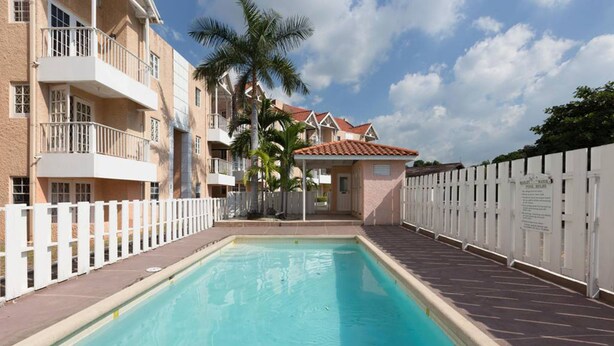 A swimming pool with a white fence, a building with balconies, and palm trees. - Kgn Most Centrally Located One Bdrm II (Kingston)