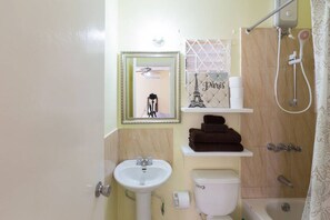 Casa de banho com lavatório, sanita e chuveiro. Há uma estante com toalhas e um objeto decorativo acima da sanita.