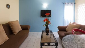 A living room with two brown sofas, a glass coffee table with a vase of red flowers, a flat-screen TV on the wall, and a window with a white curtain. - Kgn Most Centrally Located One Bdrm II (Kingston)