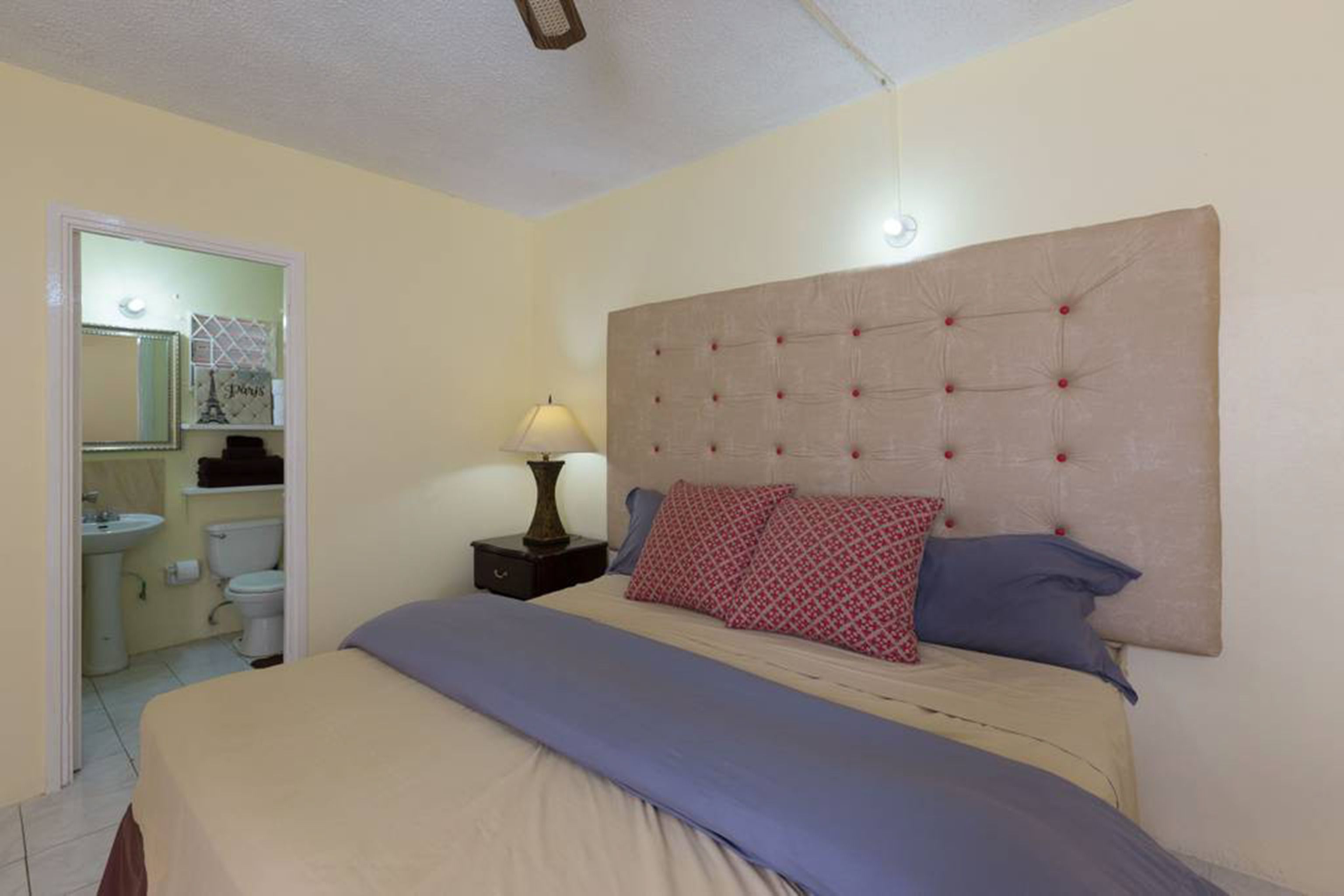 A bedroom with a large bed, a headboard, a nightstand with a lamp, and a bathroom visible through an open door.