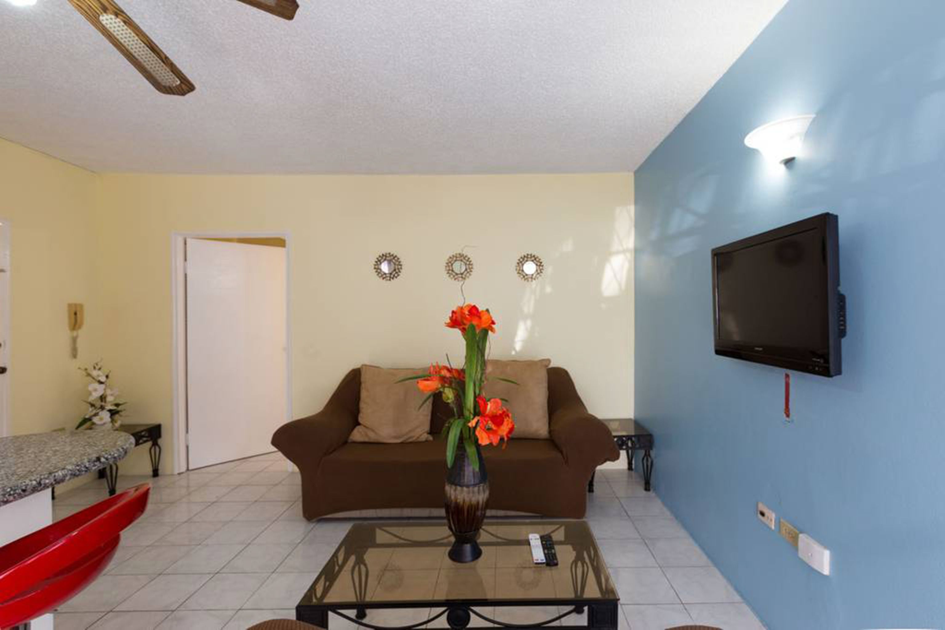 A living room with a brown sofa, a glass coffee table, a vase with flowers, a television on the wall, and a door in the background.