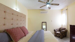 A bedroom with a tufted headboard, a bed with patterned pillows, a ceiling fan, an air conditioning unit, a mirror, a patterned armchair, and a dresser. - Kgn Most Centrally Located One Bdrm II (Kingston)