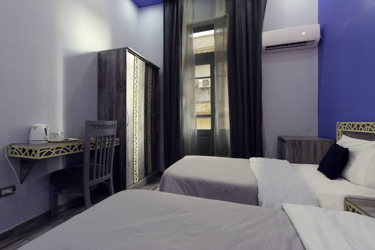 A hotel room with two beds, a desk with a chair, a wardrobe, and an air conditioning unit.