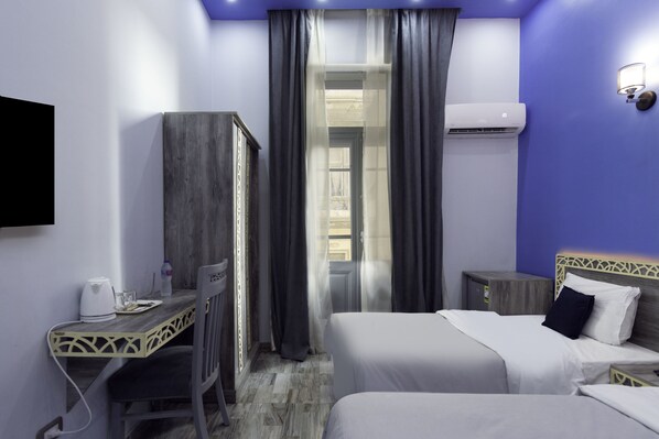 Ein Hotelzimmer mit zwei Betten, einem Schreibtisch, einem Sessel, einem Fernseher, einer Klimaanlage und einem Fenster mit Vorhängen.
