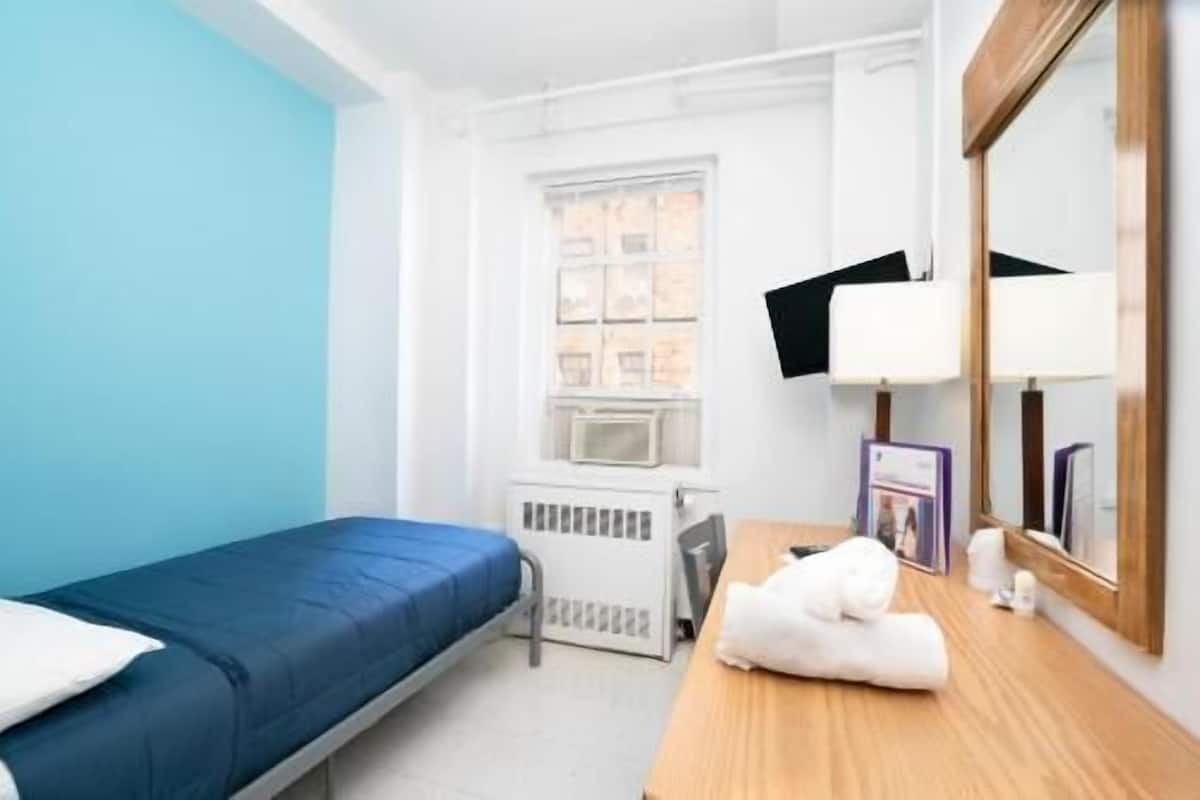 A single bed with a blue mattress, a wooden bedside table with a lamp, a mirror, and a radiator.