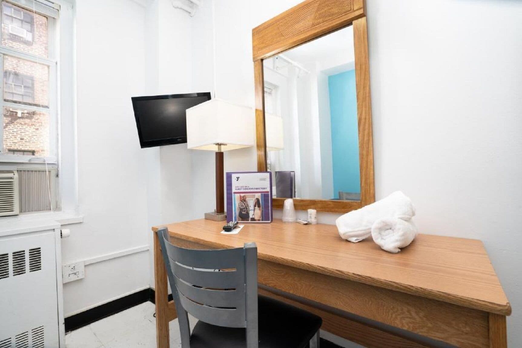 A hotel room with a wooden desk, a chair, a television mounted on the wall, a mirror, a lamp, a brochure, and a towel on the desk.
