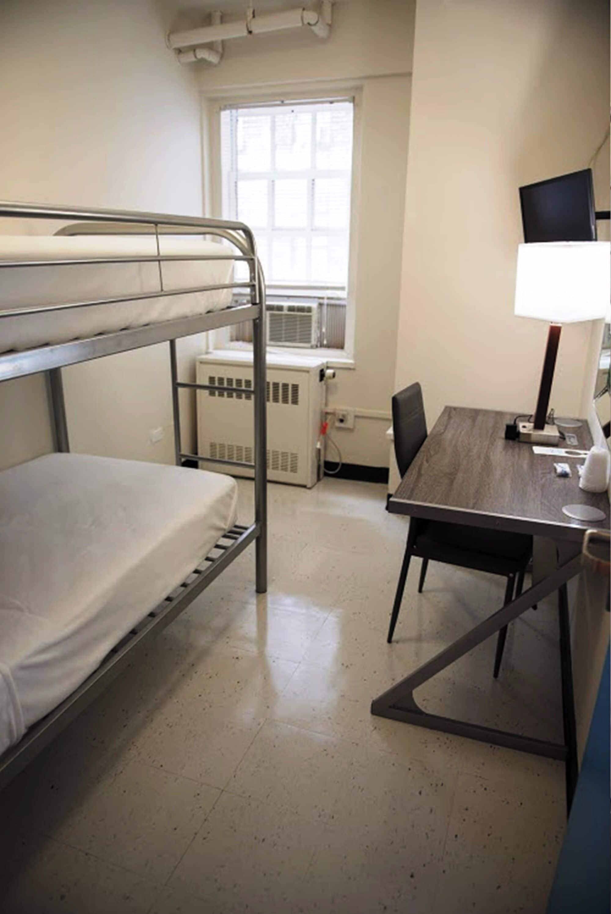 A dormitory room with bunk beds, a desk, a chair, a computer, and a window with blinds.
