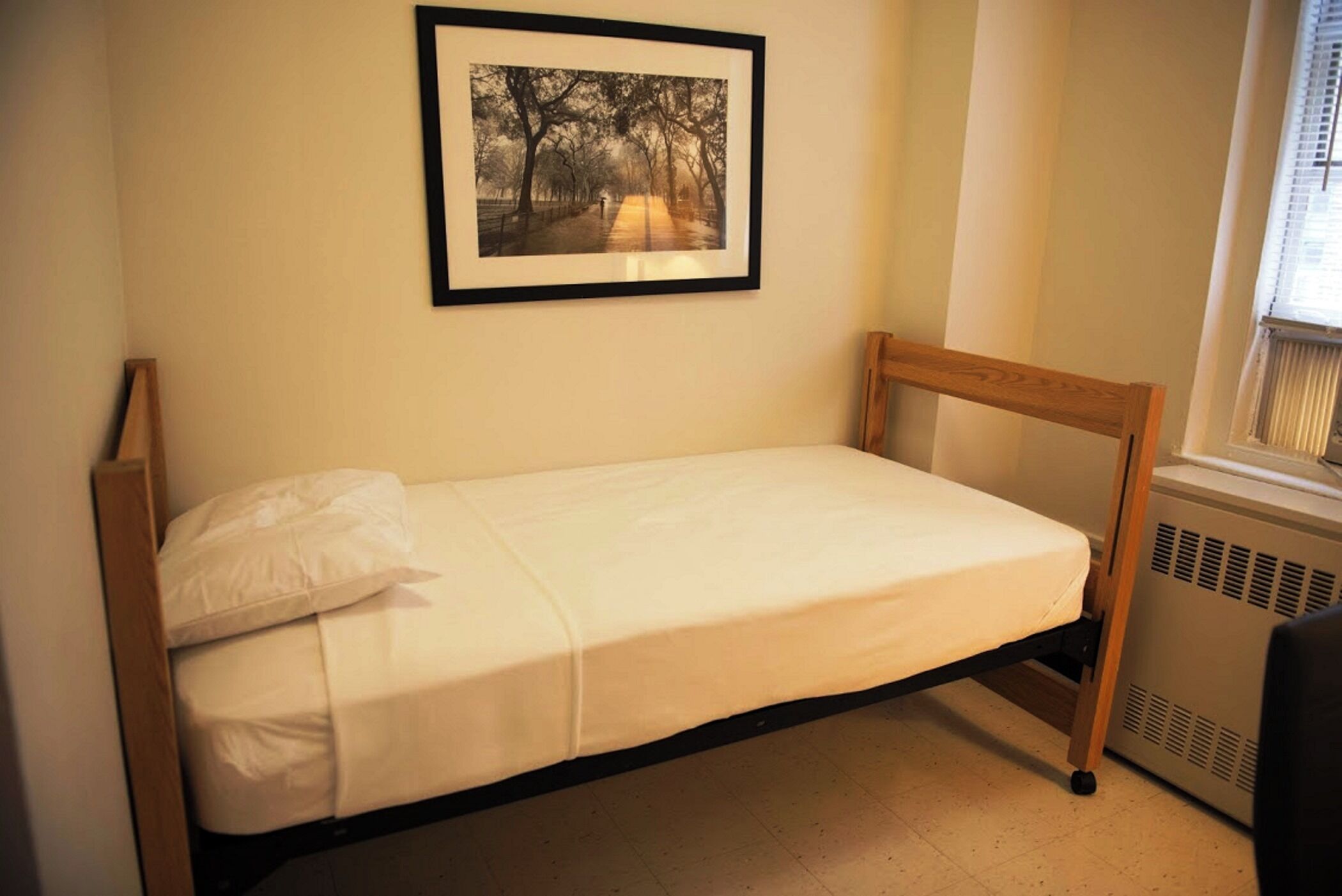 Una cama individual con estructura de madera y ropa de cama blanca en una habitación con radiador y un cuadro enmarcado en la pared.