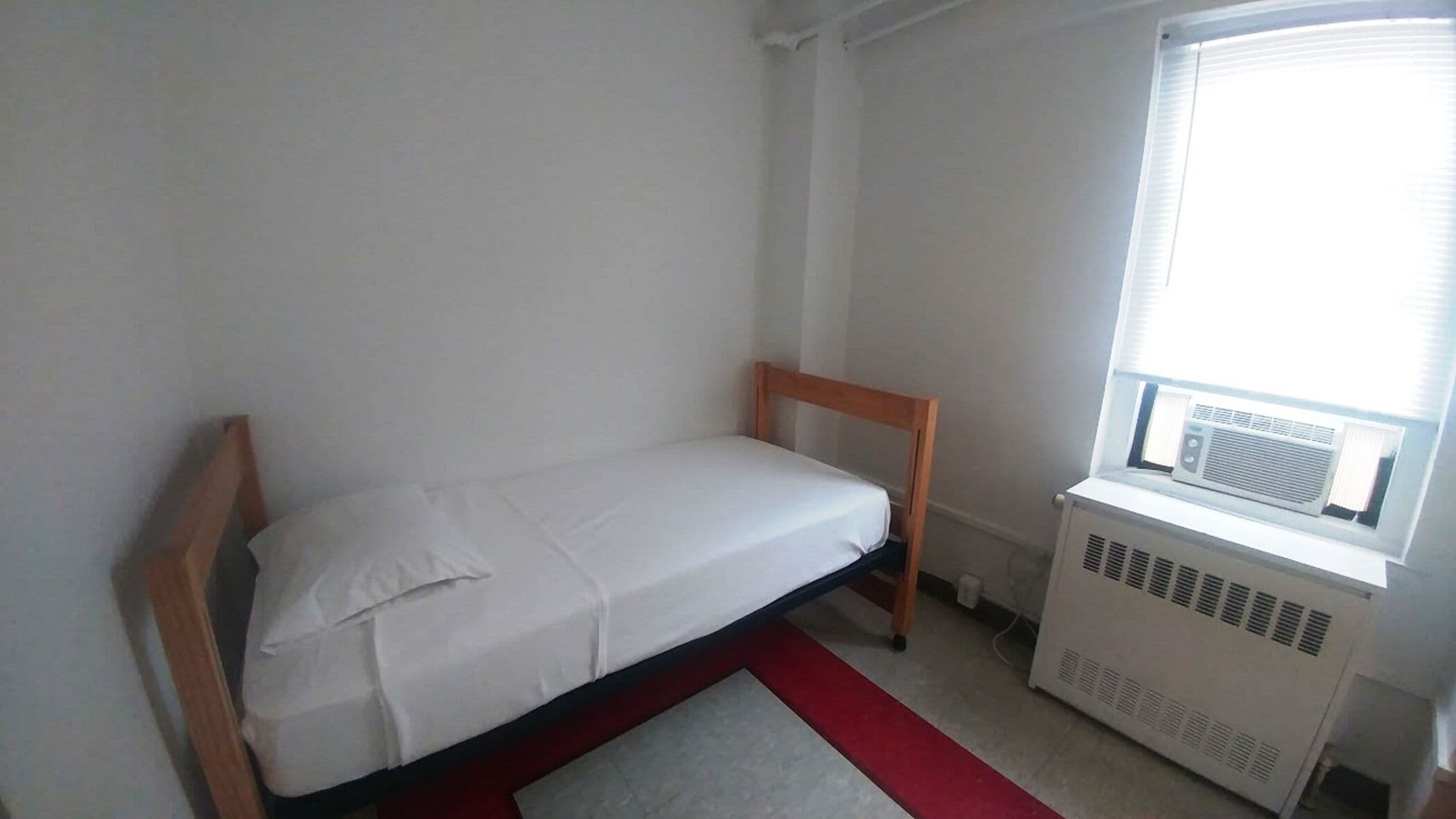 Une petite chambre avec un lit simple, une tête de lit en bois, une fenêtre avec un appareil de climatisation et un tapis rouge sur le sol.