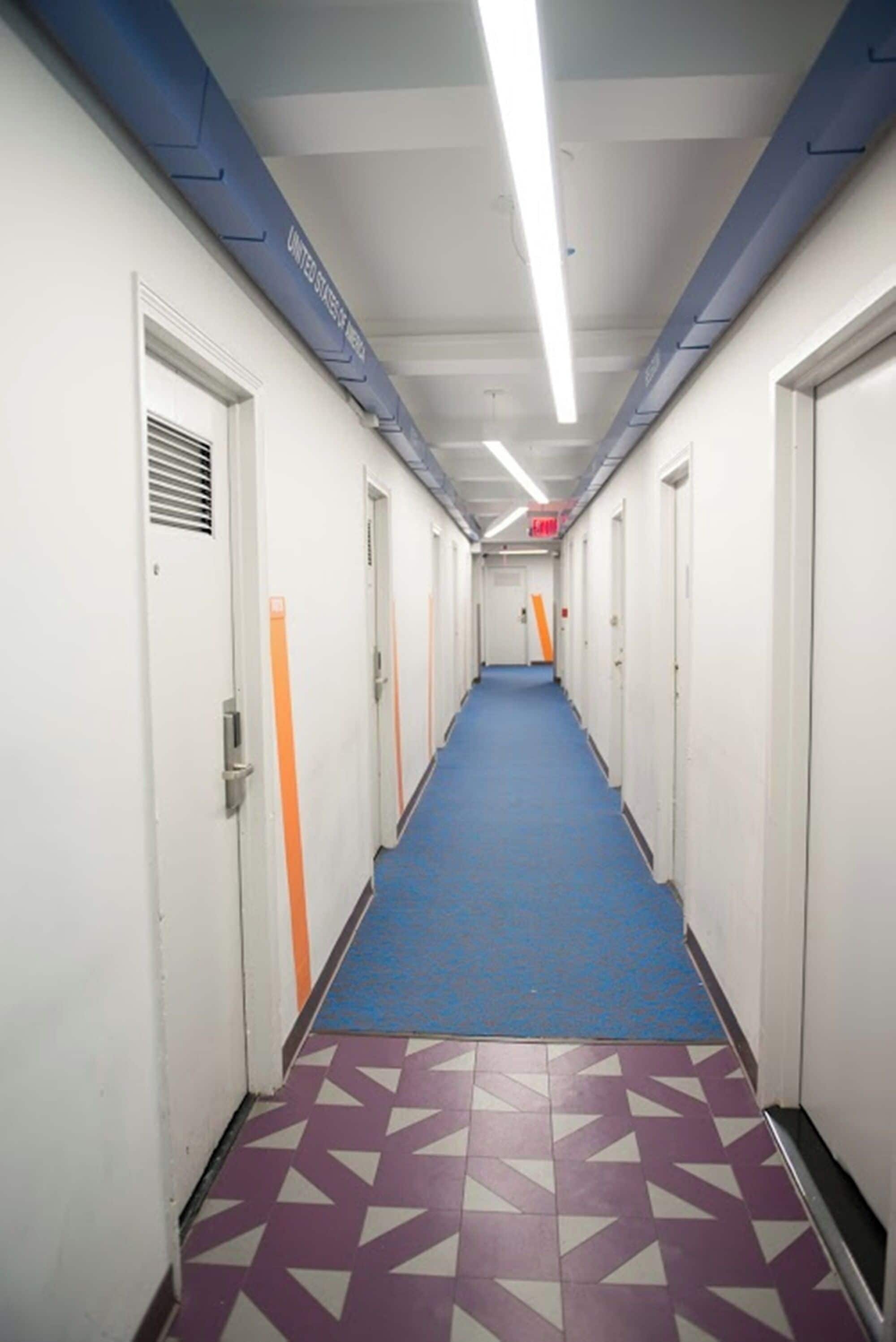 A long, narrow hallway with blue carpet and patterned floor tiles, white doors with orange stripes, and a blue ceiling with ventilation ducts.