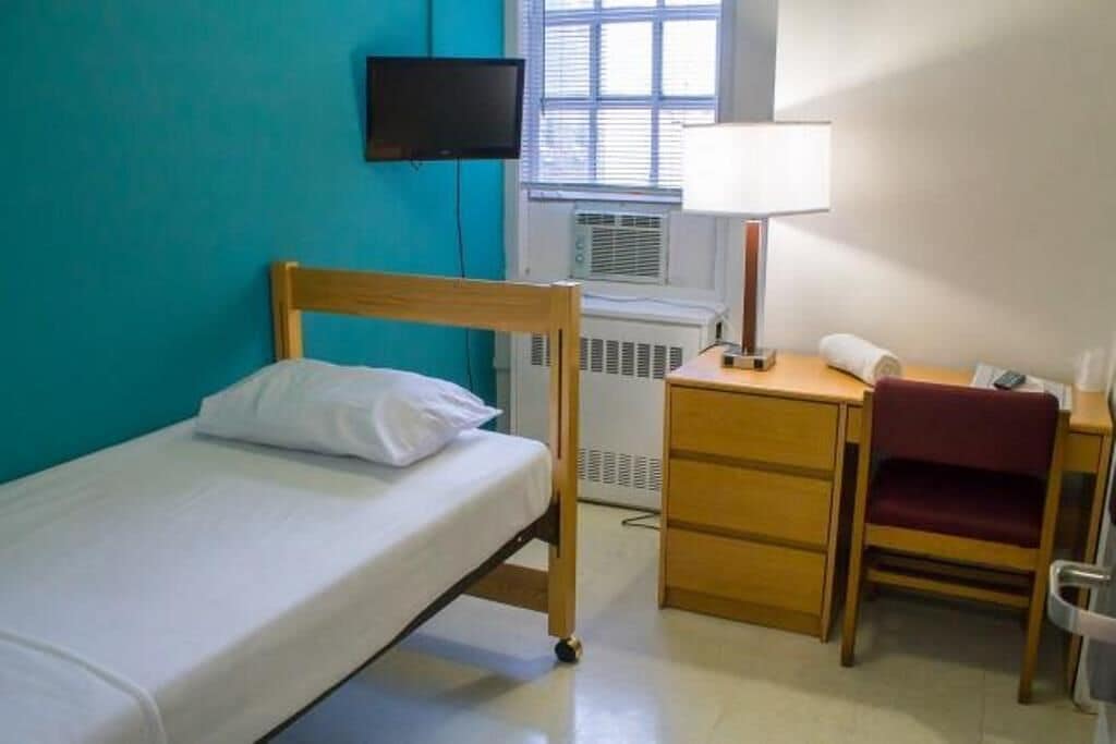 Una cama individual con cabecera de madera, un escritorio con silla, una lámpara y un televisor montado en la pared.