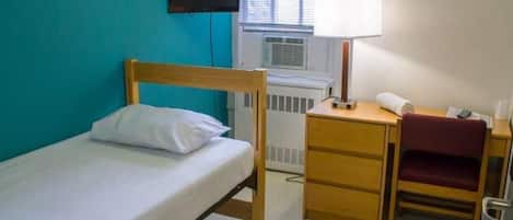 Una cama individual con cabecera de madera, un escritorio con silla, una lámpara y un televisor montado en la pared.