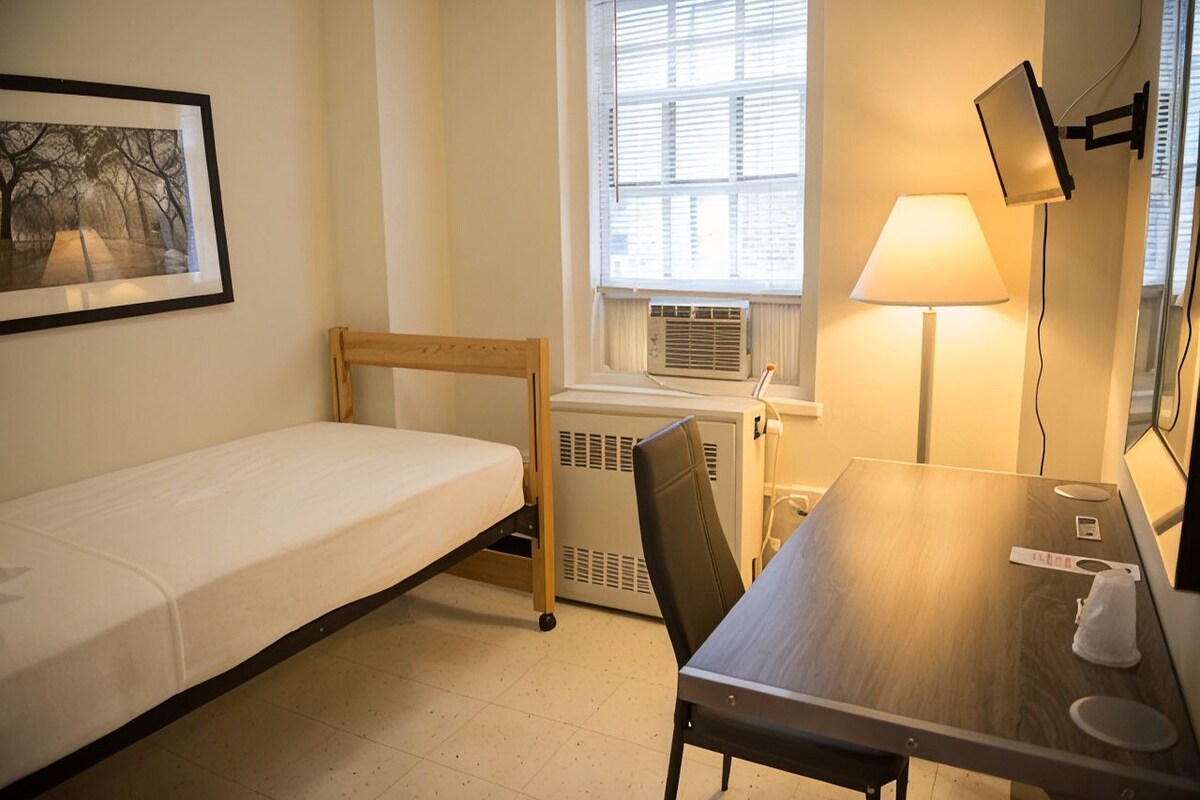 A small, single-bed room with a wooden headboard, a desk with a chair, a standing lamp, and a window with blinds.