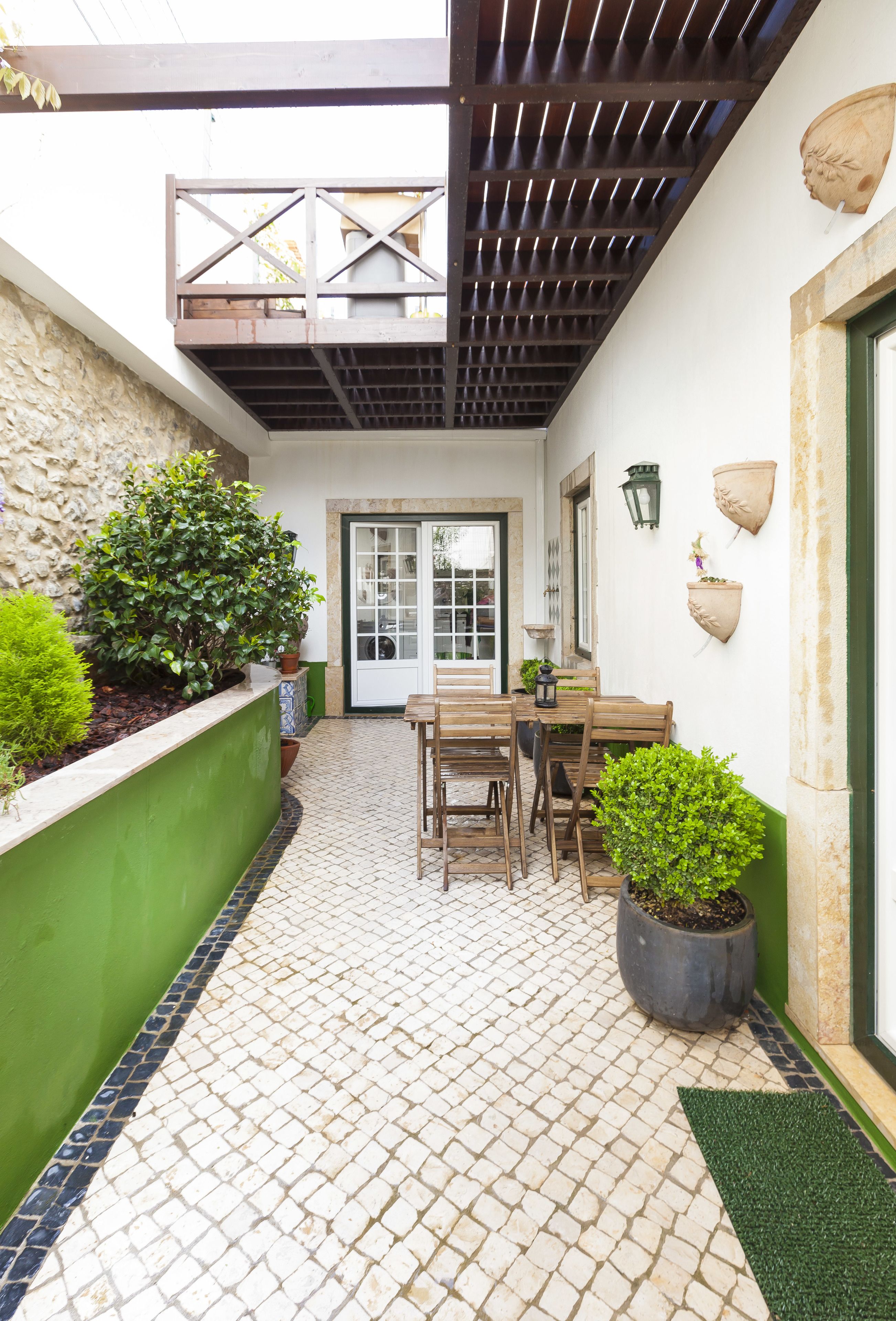 Un patio cubierto con piso de baldosas, una mesa y sillas de madera, plantas en macetas y un muro de piedra.