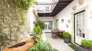 Un patio con suelo de ladrillo, una mesa y sillas de madera, plantas en macetas y un muro de piedra.