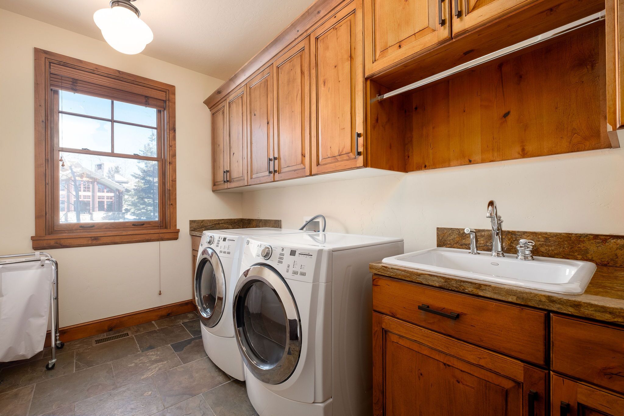 A laundry area with two front-loading washing machines, a sink with a granite countertop, wooden cabinets, and a window with a view of a house.
