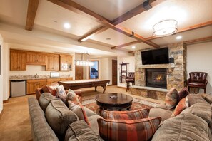 A spacious living room with a pool table, comfortable seating, a stone fireplace, and a kitchen with wooden cabinets.