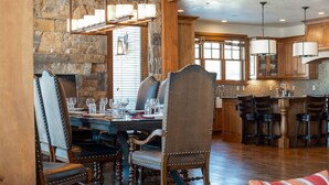 A dining area with a stone wall, wooden ceiling beams, and a chandelier. There are multiple dining tables and chairs, and a kitchen area with wooden cabinets and bar stools. - Managed by Park City Lodging-Private Home with Stunning Deer Valley Resort Views (Park City)