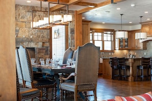 A dining area with a stone wall, wooden ceiling beams, and a chandelier. There are multiple dining tables and chairs, and a kitchen area with wooden cabinets and bar stools.