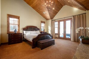 A bedroom with a wooden ceiling, a large bed, a bench, a nightstand, and a window with curtains.