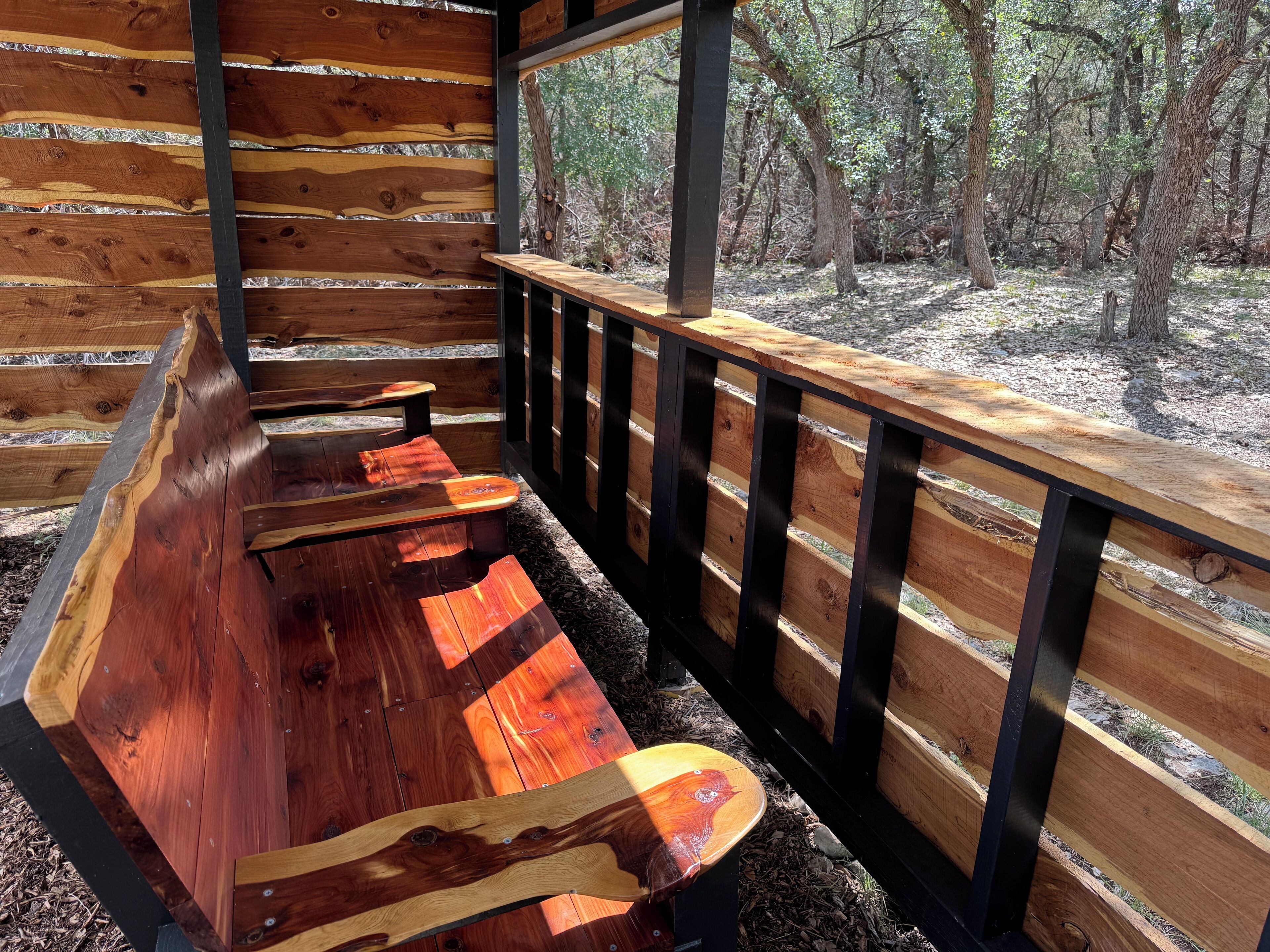 A wooden bench with a metal railing in a covered outdoor area with a view of a forest.
