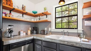 A modern kitchen with a dishwasher, a coffee maker, and a toaster. There is a window with a view of trees and a shelf with jars and a plant.