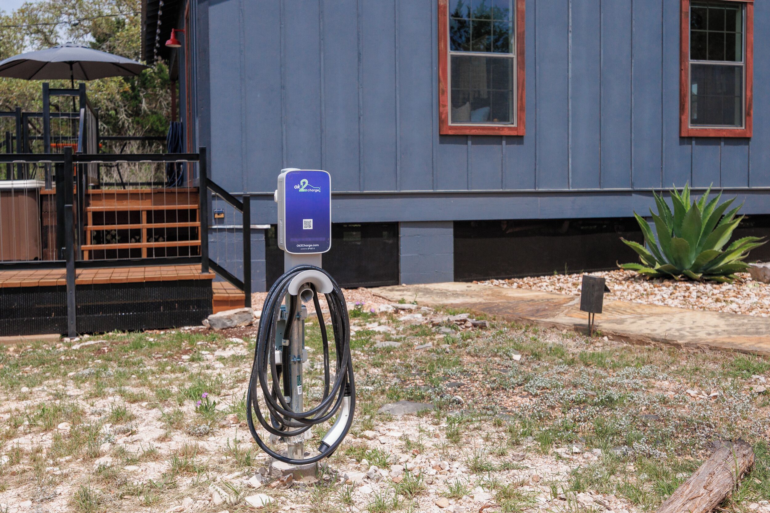 An electric vehicle charging station in front of a house with a deck and a large plant.
