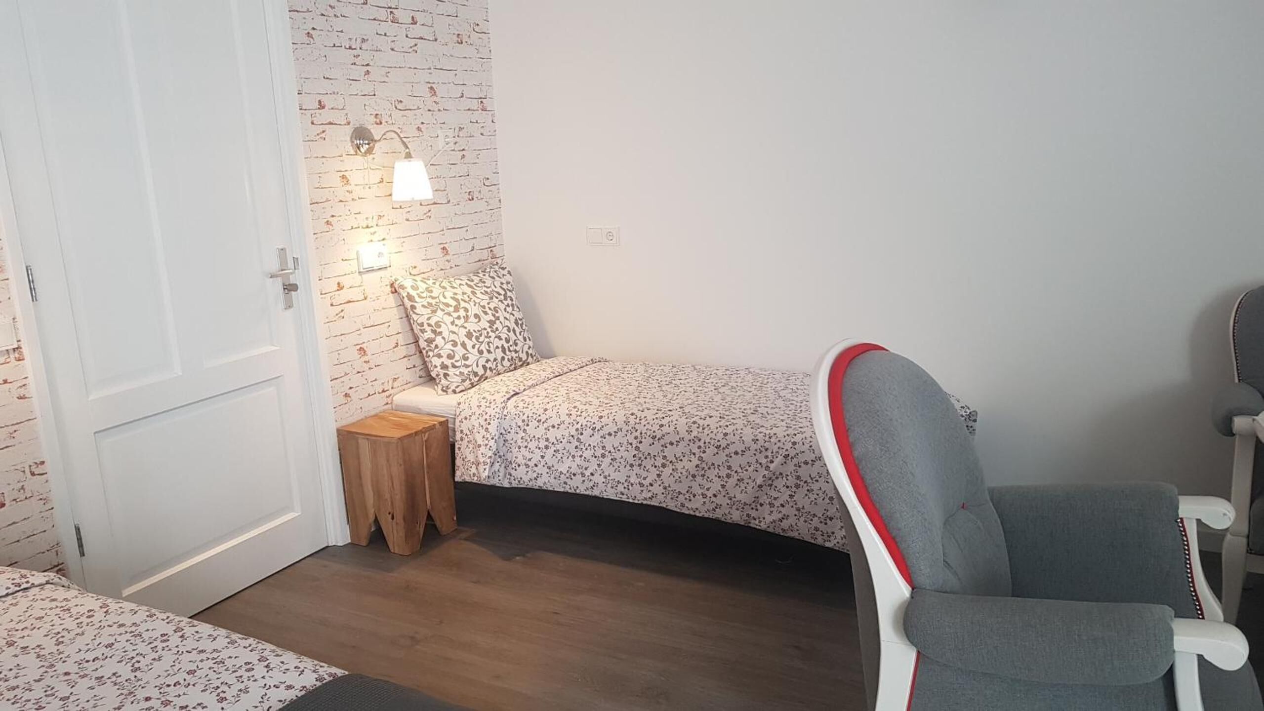 Una habitación pequeña y acogedora con una cama, una silla, un taburete de madera y una pared de ladrillo con una lámpara.