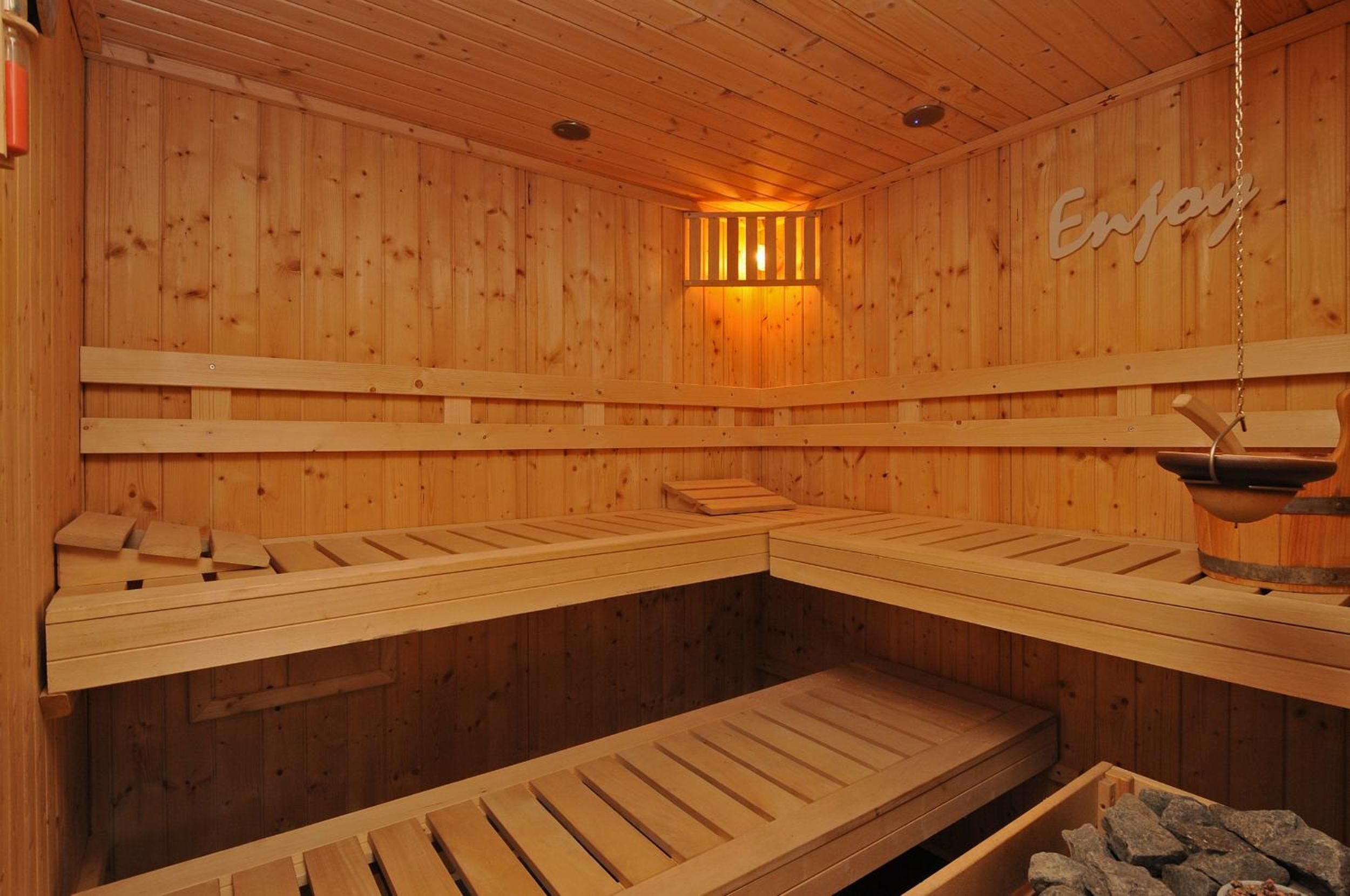 Een sauna met houten wanden en banken, een hangende waterbak en een bordje met de tekst 'Geniet'.