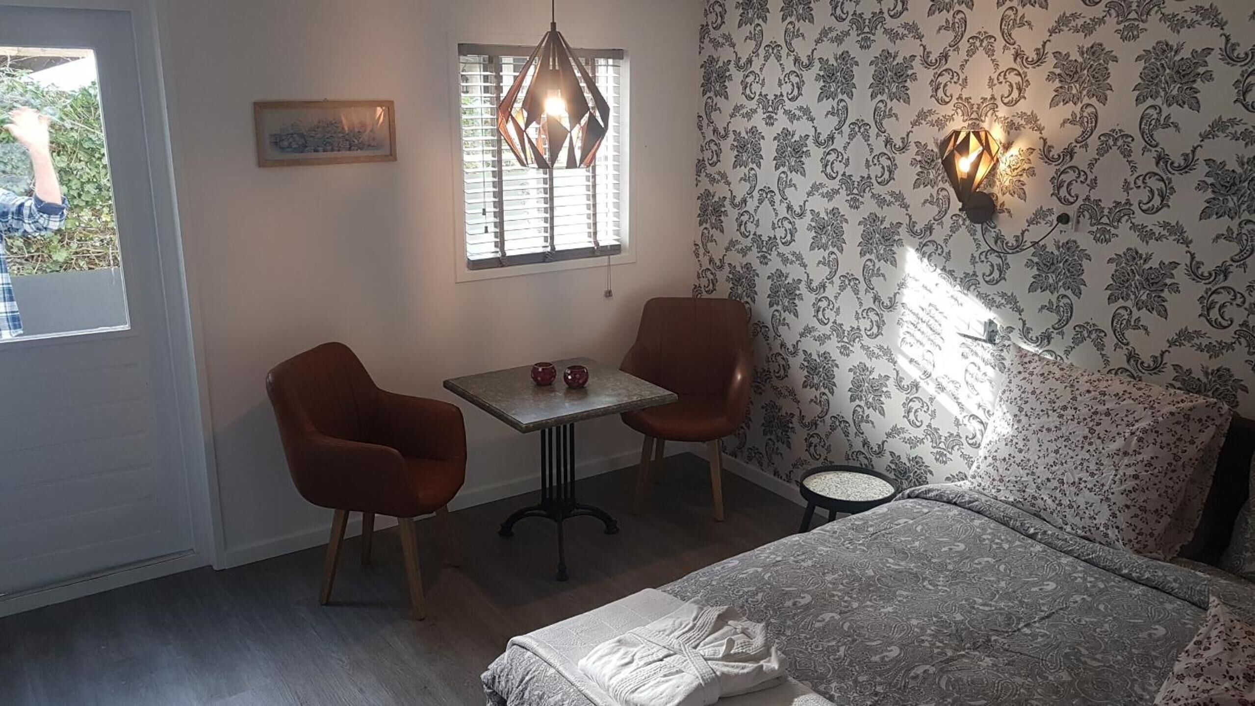 Ein Schlafzimmer mit einem Bett, zwei Stühlen, einem kleinen Tisch, einer Hängeleuchte, einem Fenster und einer Wand mit strukturierter Tapete.