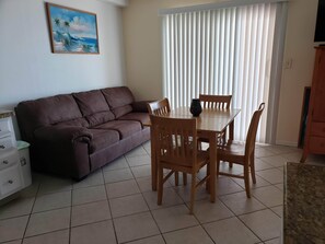 A living room with a brown sofa, wooden dining table, and chairs. There is a painting on the wall and a window with blinds.