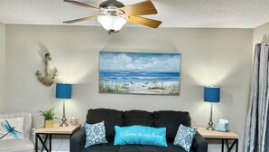 A living room with a ceiling fan, a painting of a beach, a black sofa with patterned cushions, a coffee table, and two lamps on side tables.