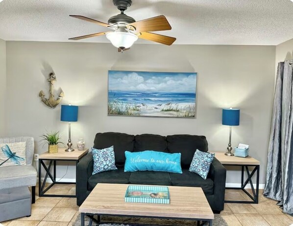 A living room with a ceiling fan, a painting of a beach, a black sofa with patterned cushions, a coffee table, and two lamps on side tables.