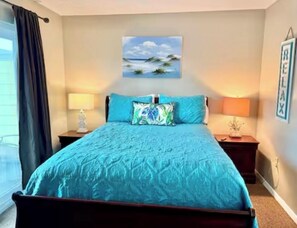 A bedroom with a bed, two nightstands, a painting of a beach scene, a window with curtains, and a wall with a framed word. - Gulf View, Sandy Beach Across Street, Near Pier Park (Panama City Beach)