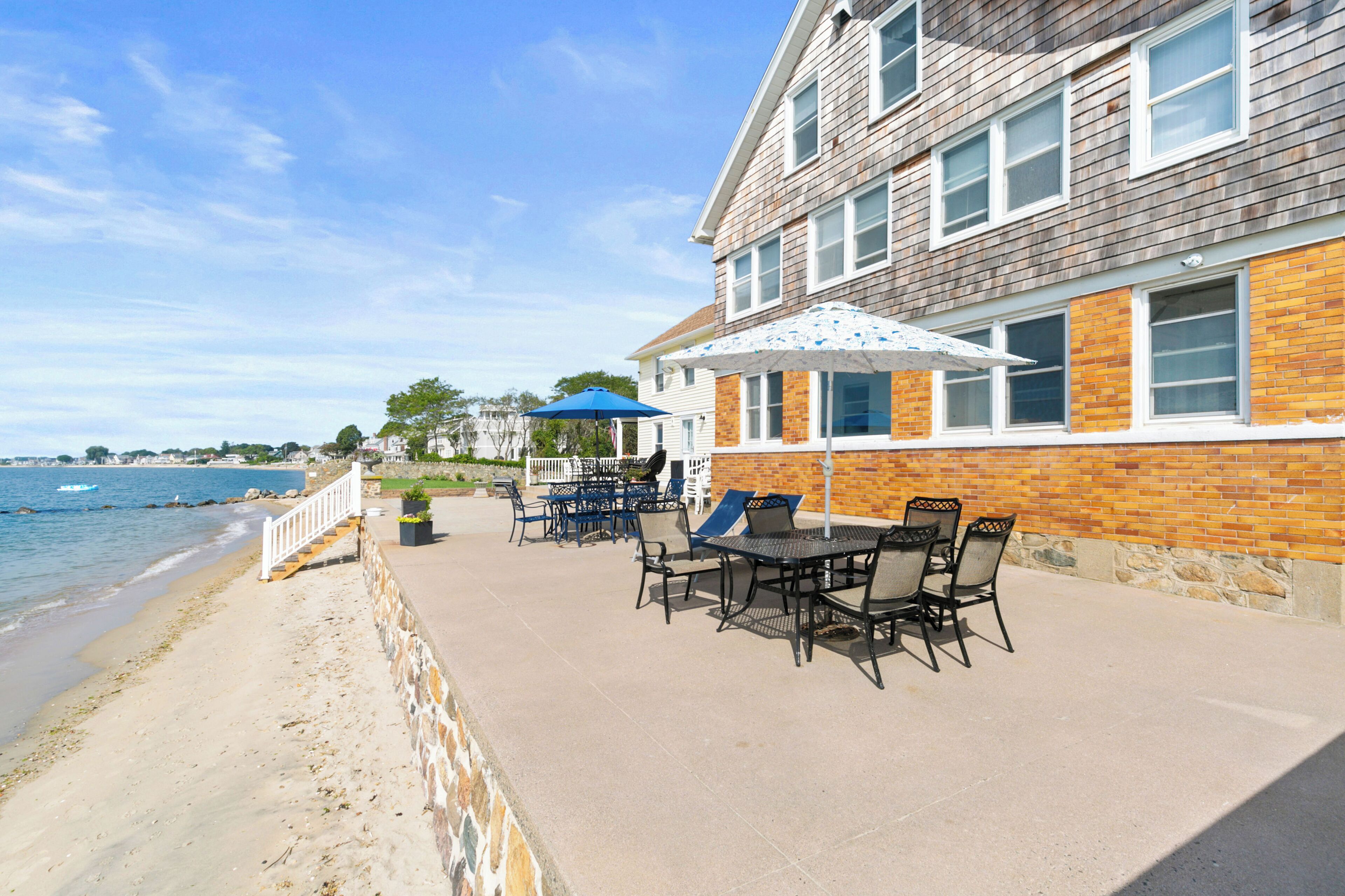 A beachfront patio with outdoor furniture and umbrellas, a stone wall, and a view of the sea.