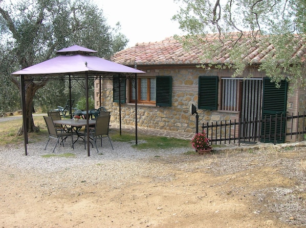 Una casa de piedra con tejado de baldosas, jardín vallado y un espacio exterior cubierto con sillas y una mesa.