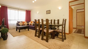 A living room with a sofa, coffee table, dining table, and chairs. There is a TV, a plant, and a door leading to another room. - City Airport Apartment Nairobi (Syokimau)