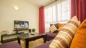 A living room with a television, a sofa, a coffee table, and a window with curtains. - City Airport Apartment Nairobi (Syokimau)