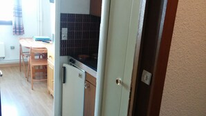 Een kleine keuken met een compacte koelkast, een magnetron en een spoelbak. Er is een eethoek met een tafel en stoelen. De keuken heeft een betegelde spatwand en houten keukenkastjes.