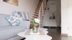 Una sala de estar con un sofá gris, almohadones estampados, una mesa de centro blanca con un jarrón de flores blancas, y una escalera que lleva a una zona de cocina.