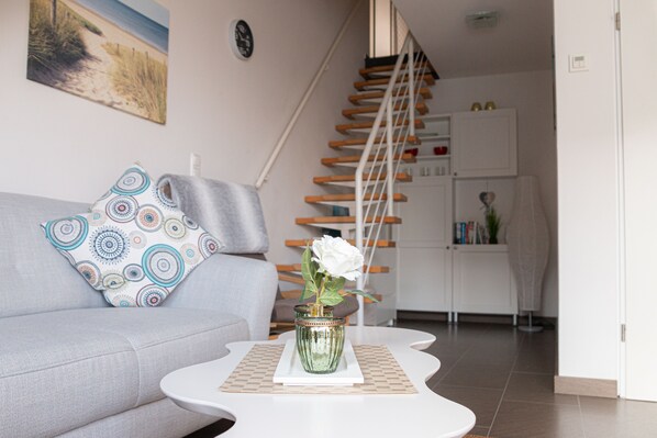 A living room with a grey sofa, patterned pillows, a white coffee table with a vase of white flowers, and a staircase leading to a kitchen area.
